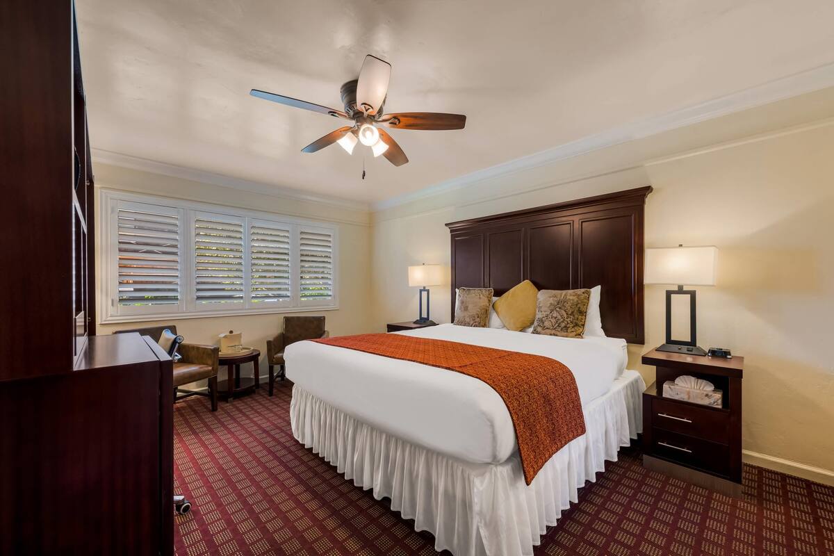 A hotel room with a large bed, a ceiling fan, a nightstand with a lamp, a wooden headboard, and a window with blinds.