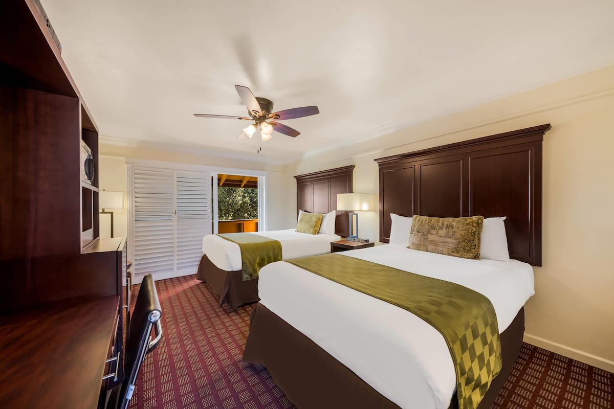 A hotel room with two beds, a ceiling fan, a nightstand, a desk, and a window with blinds.