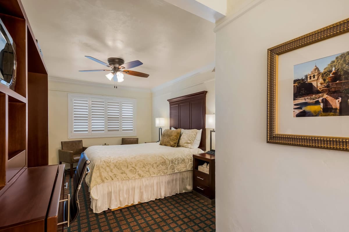 A hotel room with a bed, a desk, a chair, a ceiling fan, a window with blinds, a nightstand, a lamp, and a framed picture on the wall.