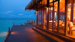 Una terraza de madera que conduce a un área de estar con ventanales y asientos, con vista al mar tranquilo.