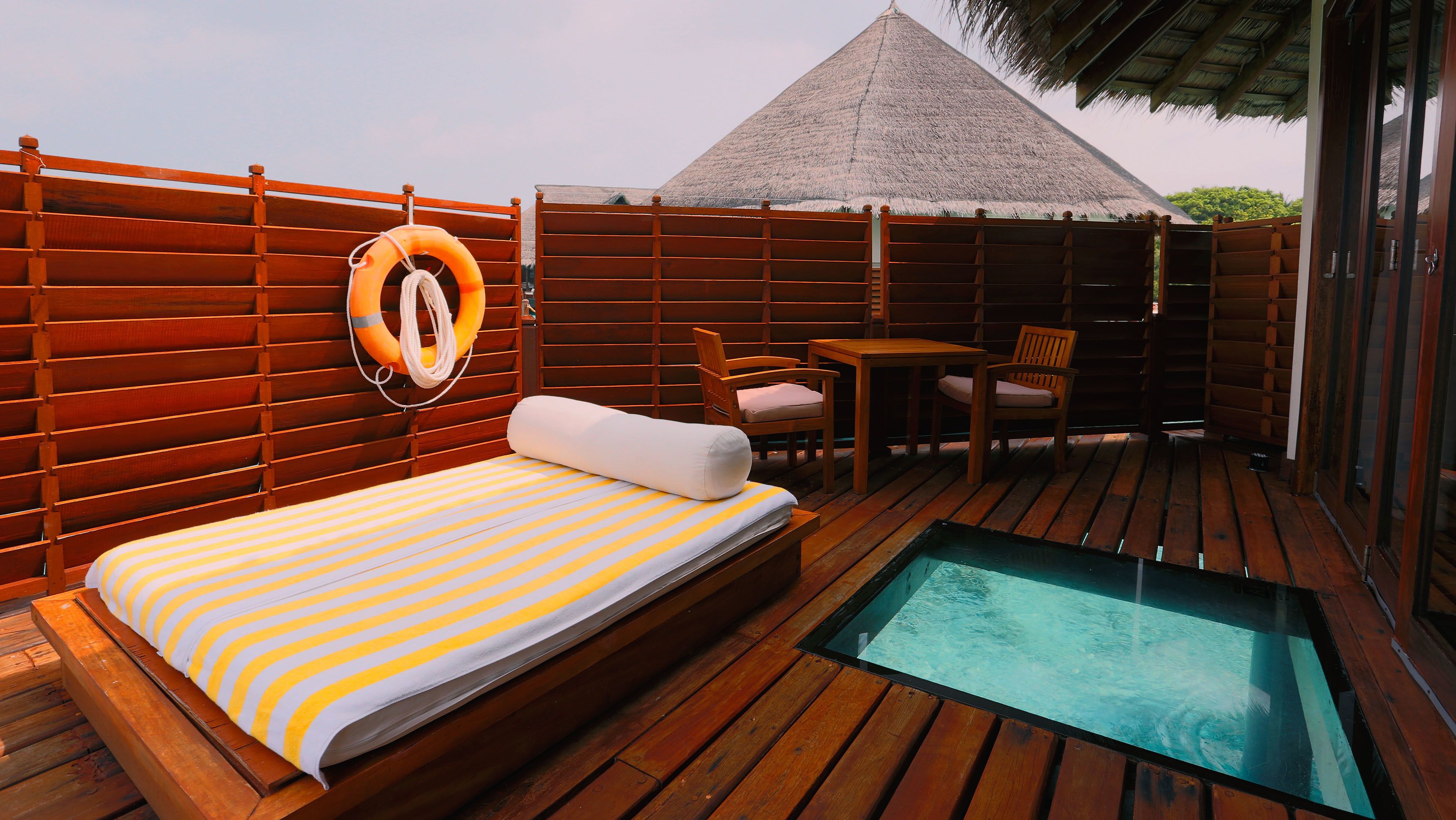 A wooden deck with a bed, a small table, two chairs, a pool, and a lifebuoy.
