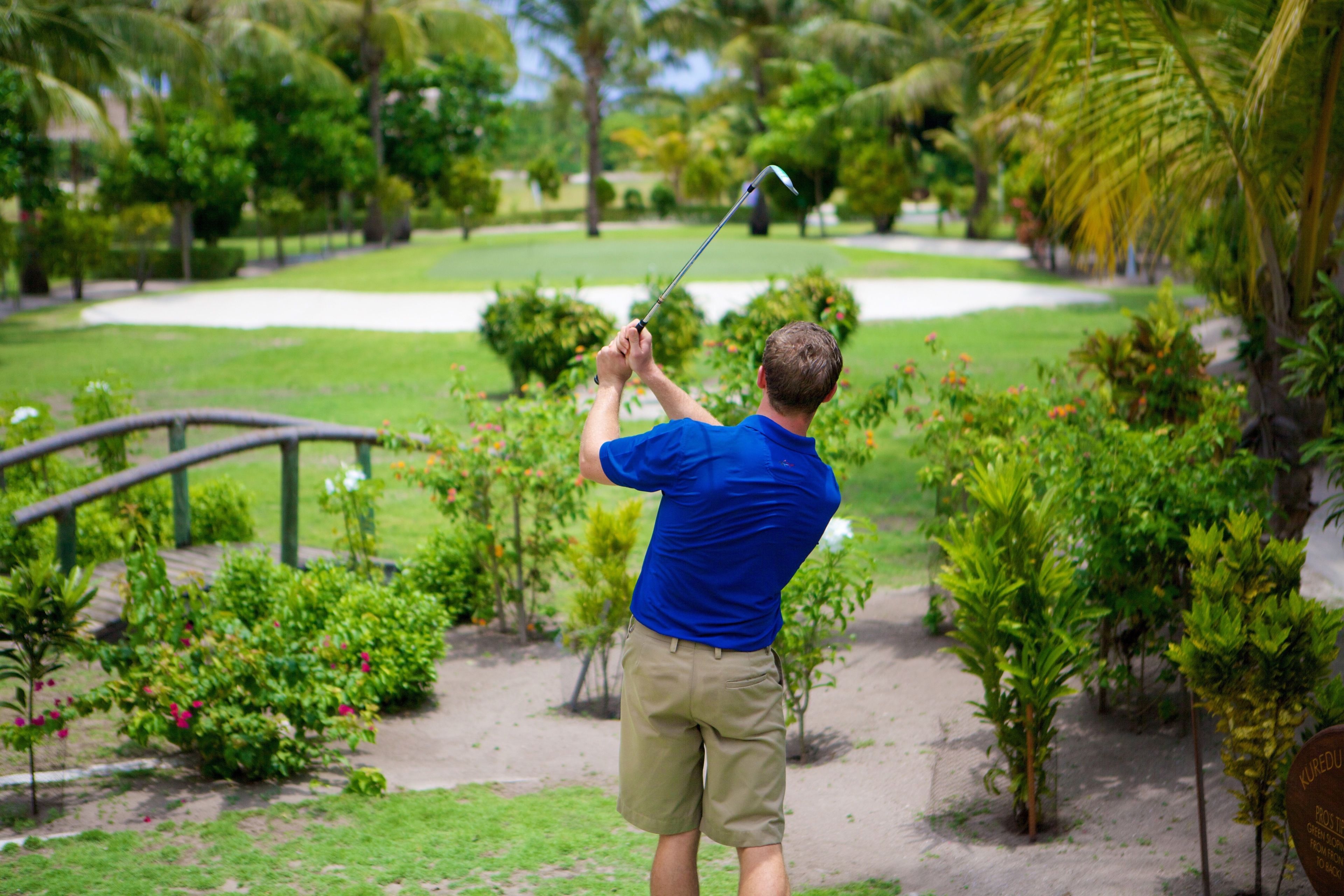 A man in a blue shirt and khaki shorts holding a golf club, standing on a golf course with a bridge and lush greenery in the background.