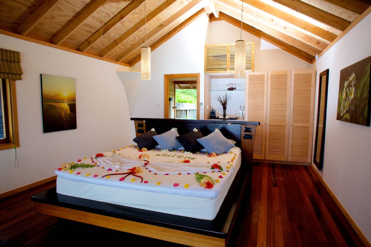 A bedroom with a wooden ceiling, a bed with a floral arrangement, a painting on the wall, and a window with blinds.