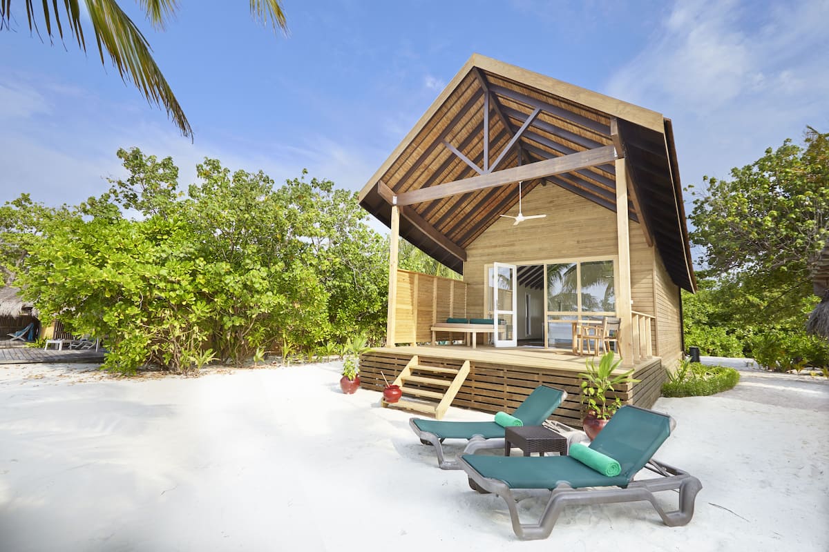 A wooden beach house with a deck, two lounge chairs, and a glass door entrance.