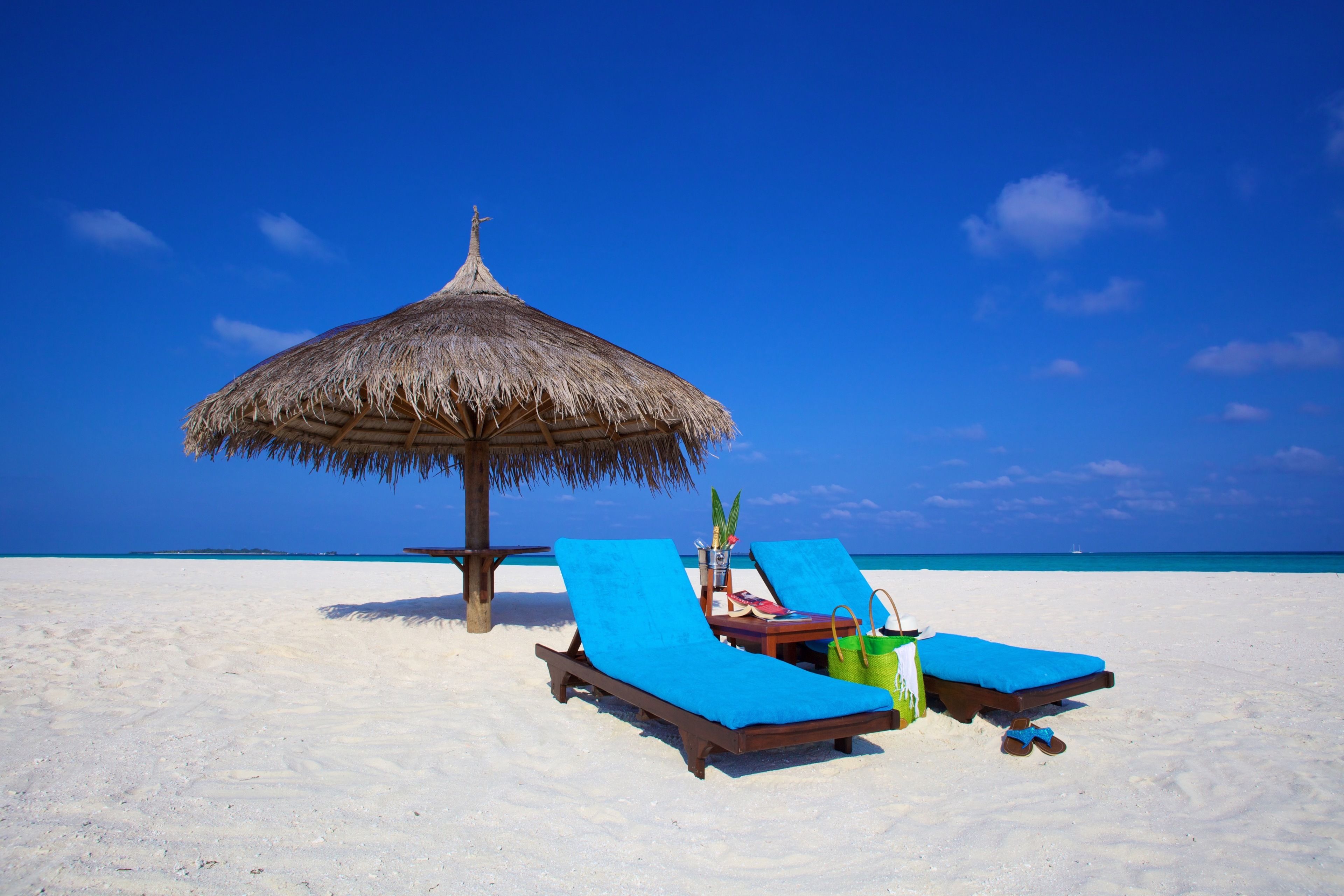 A beach with two lounge chairs, a thatched umbrella, and a small table with a plant and a bag.