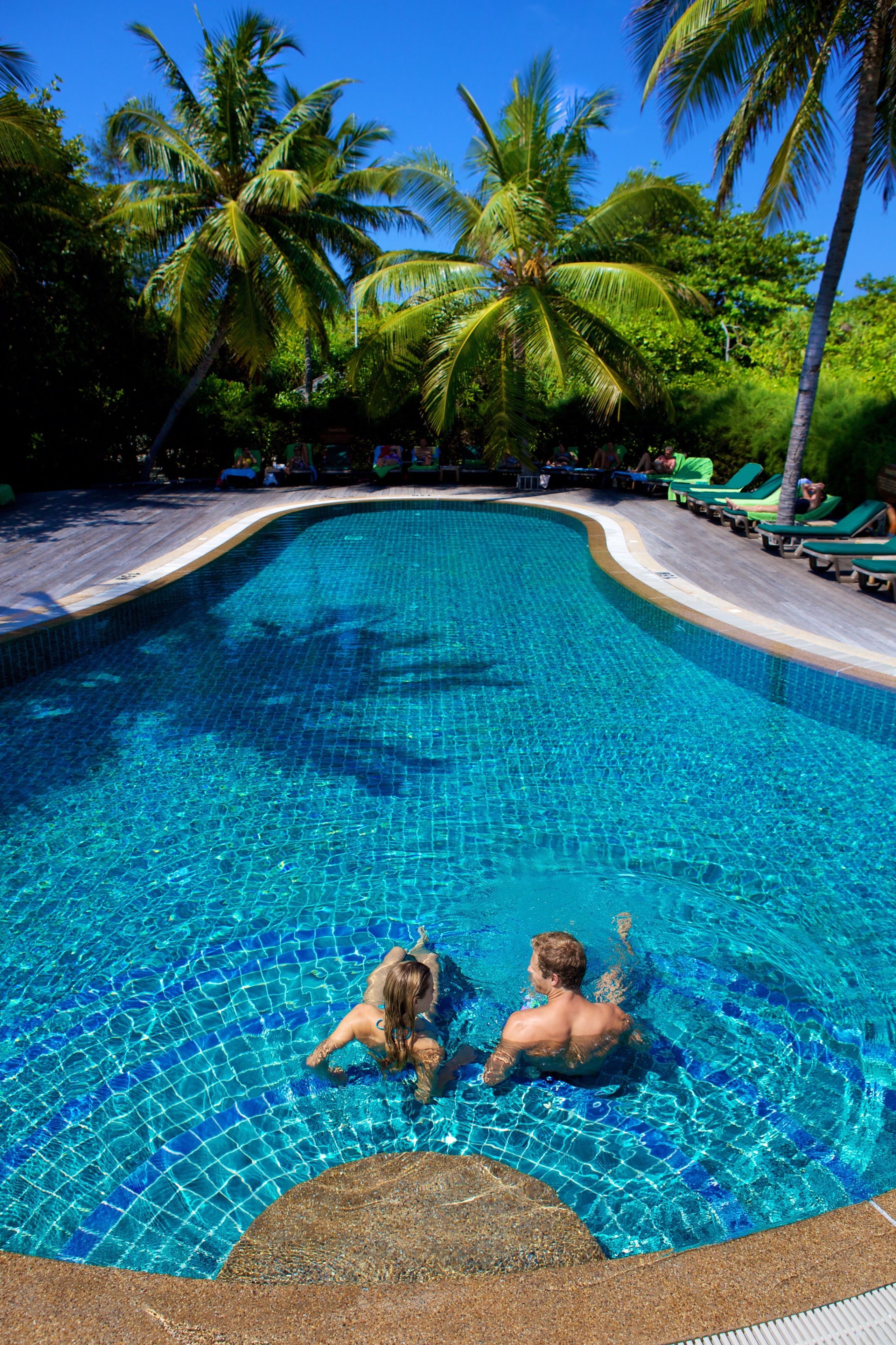 A swimming pool with clear blue water, surrounded by palm trees and lounge chairs.