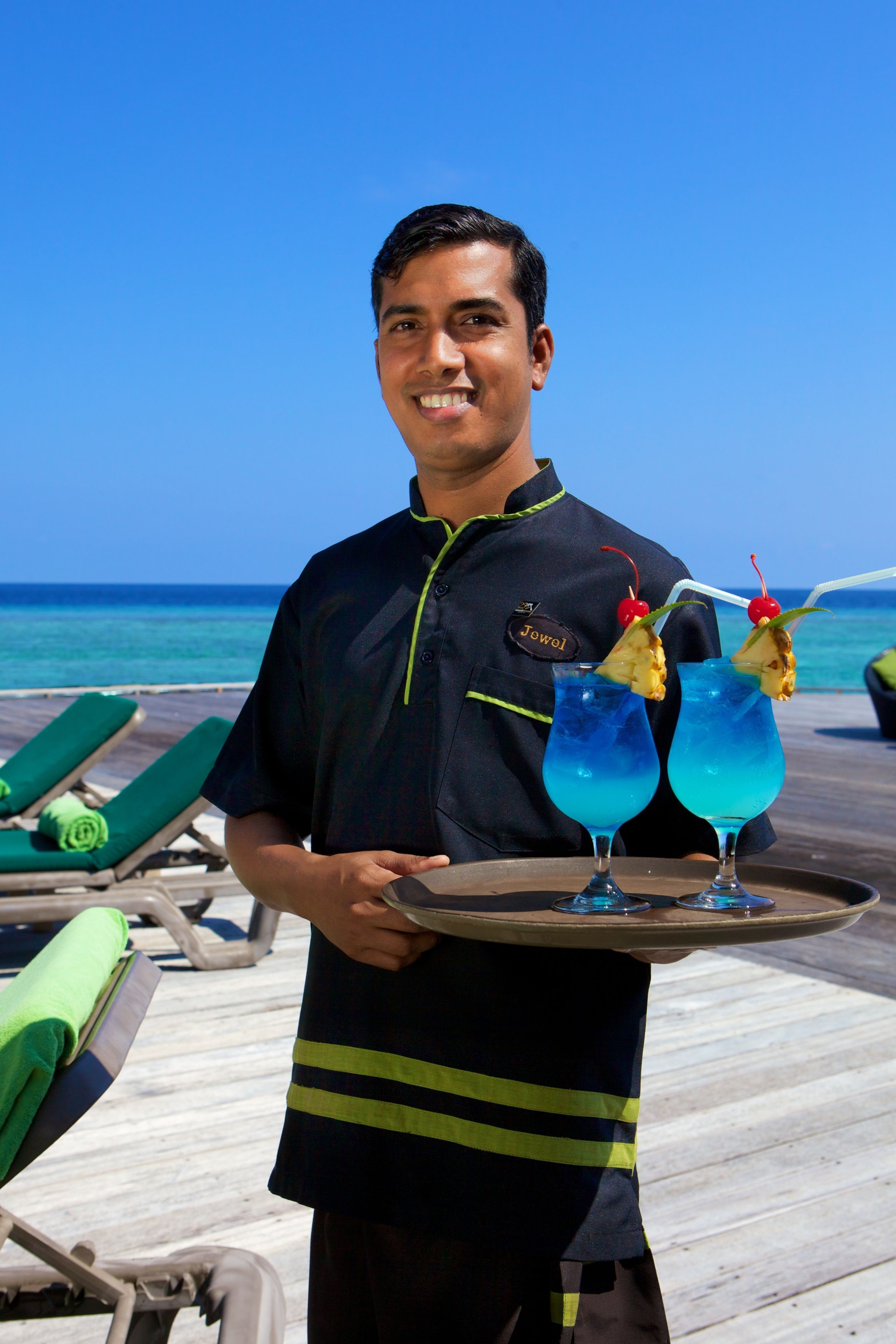 Eine Person hält ein Tablett mit zwei blauen Cocktails und einer Kirsche, stehend auf einem Holzdeck mit grünen Liegestühlen und blauem Himmel im Hintergrund.