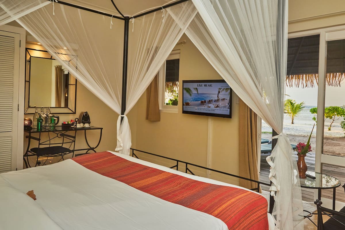 A hotel room with a bed, a TV mounted on the wall, a desk with a chair, and a view of the beach through a glass door.