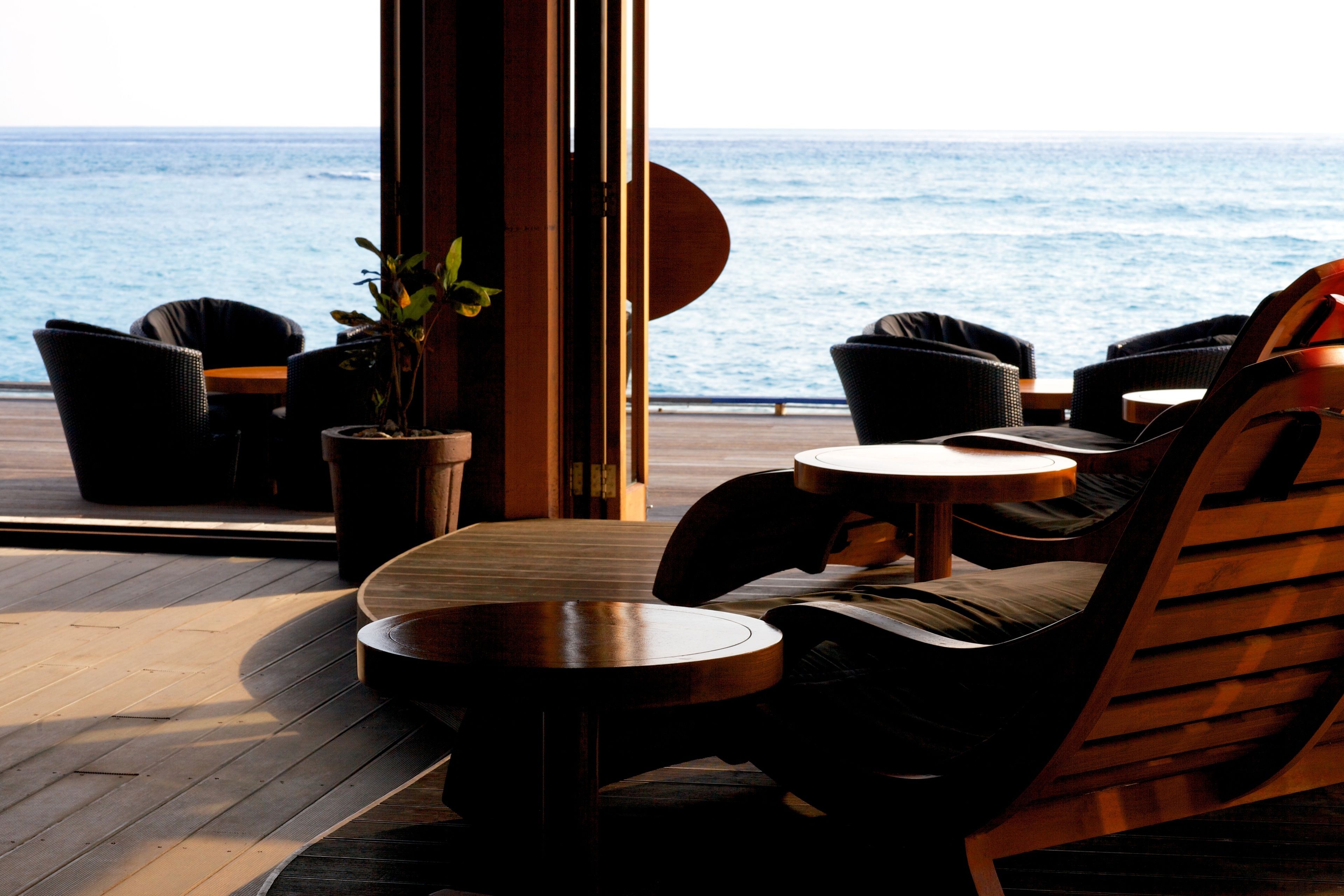 A deck with wooden flooring, round tables, and black chairs with cushions. A potted plant is visible.