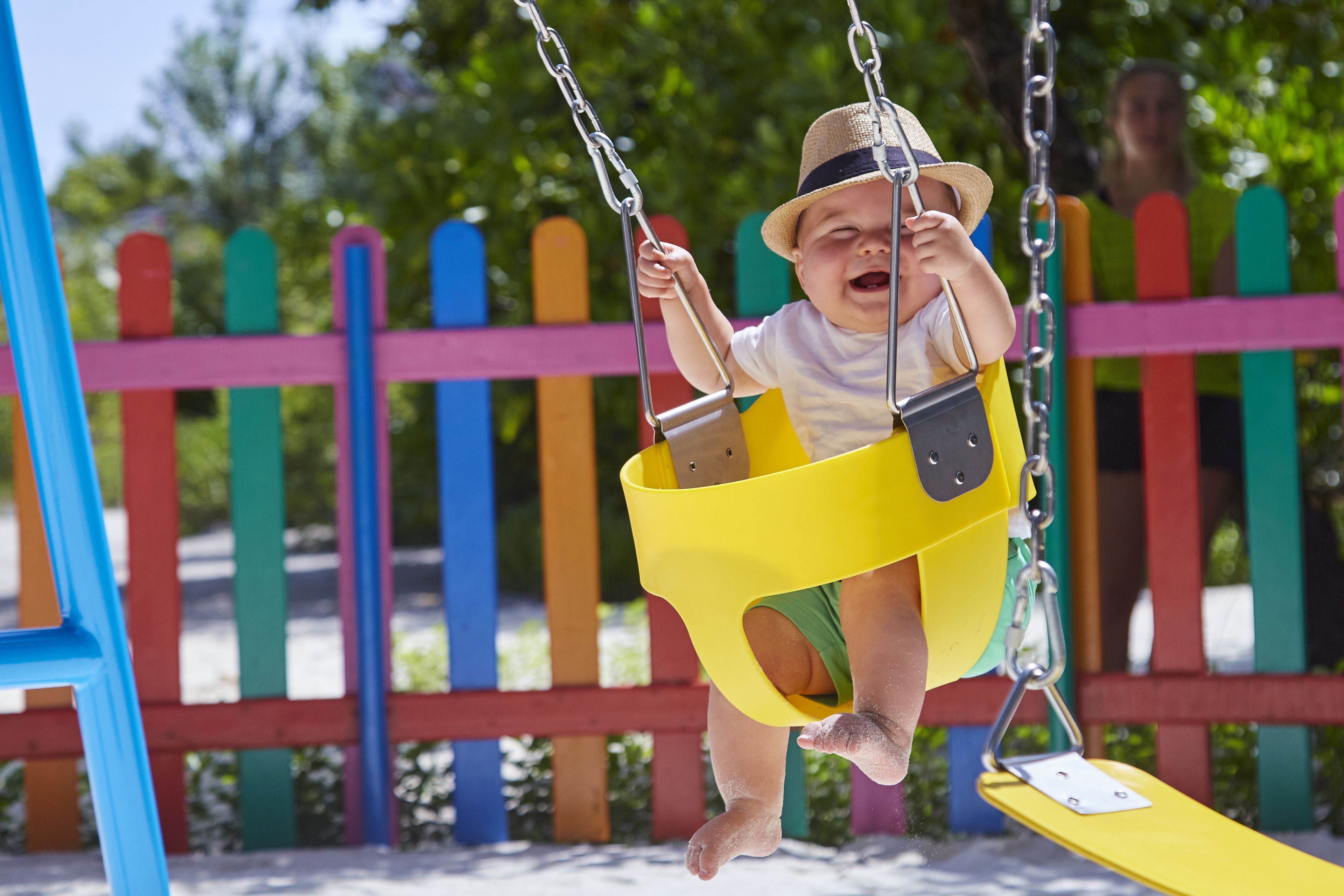 Ein Kind schwingt auf einer gelben Schaukel auf einem Spielplatz, im Hintergrund ist ein farbenfroher Zaun zu sehen.