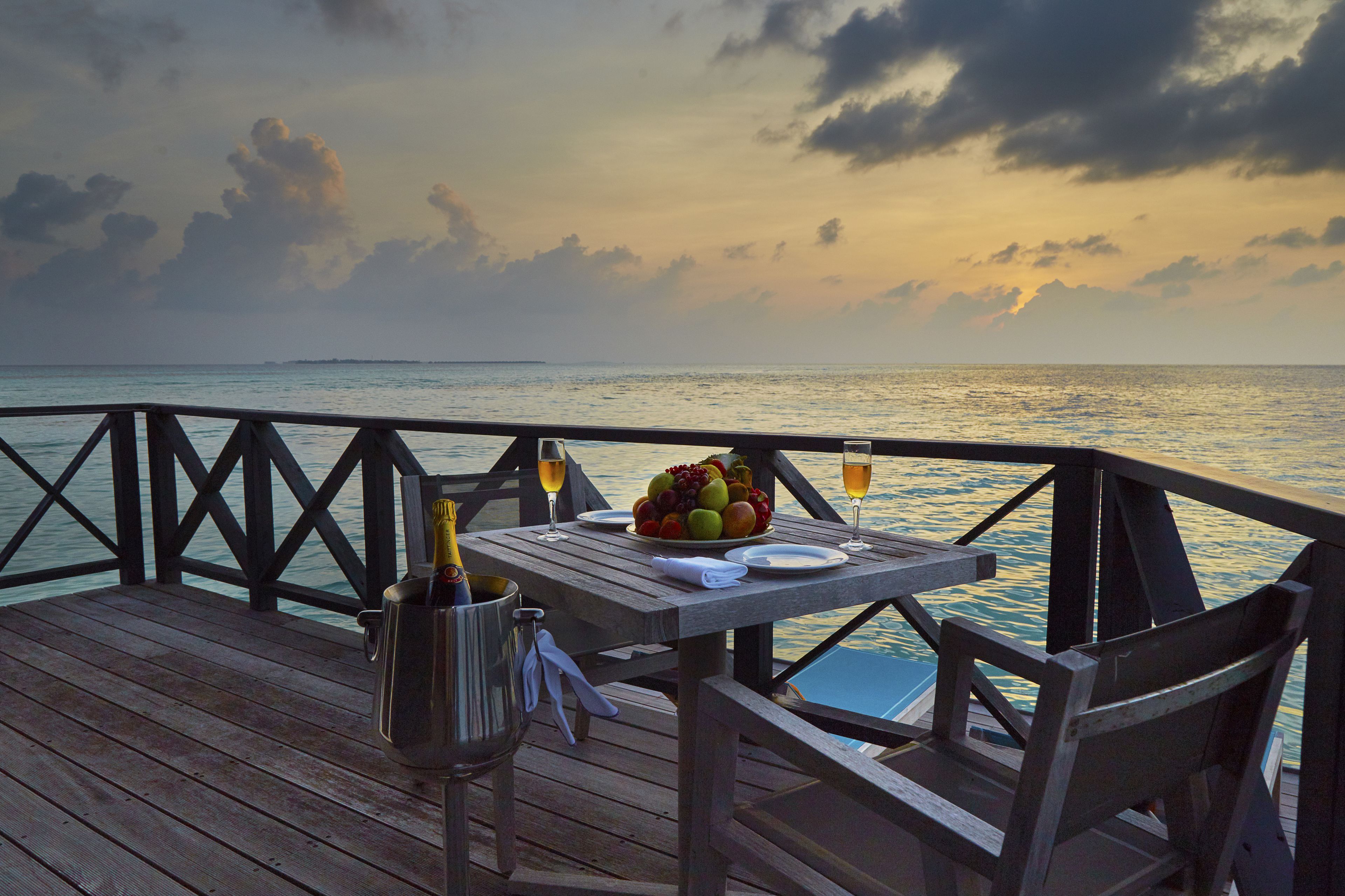 A wooden deck with a table set for two, featuring a fruit bowl, plates, glasses, and a bottle of champagne.