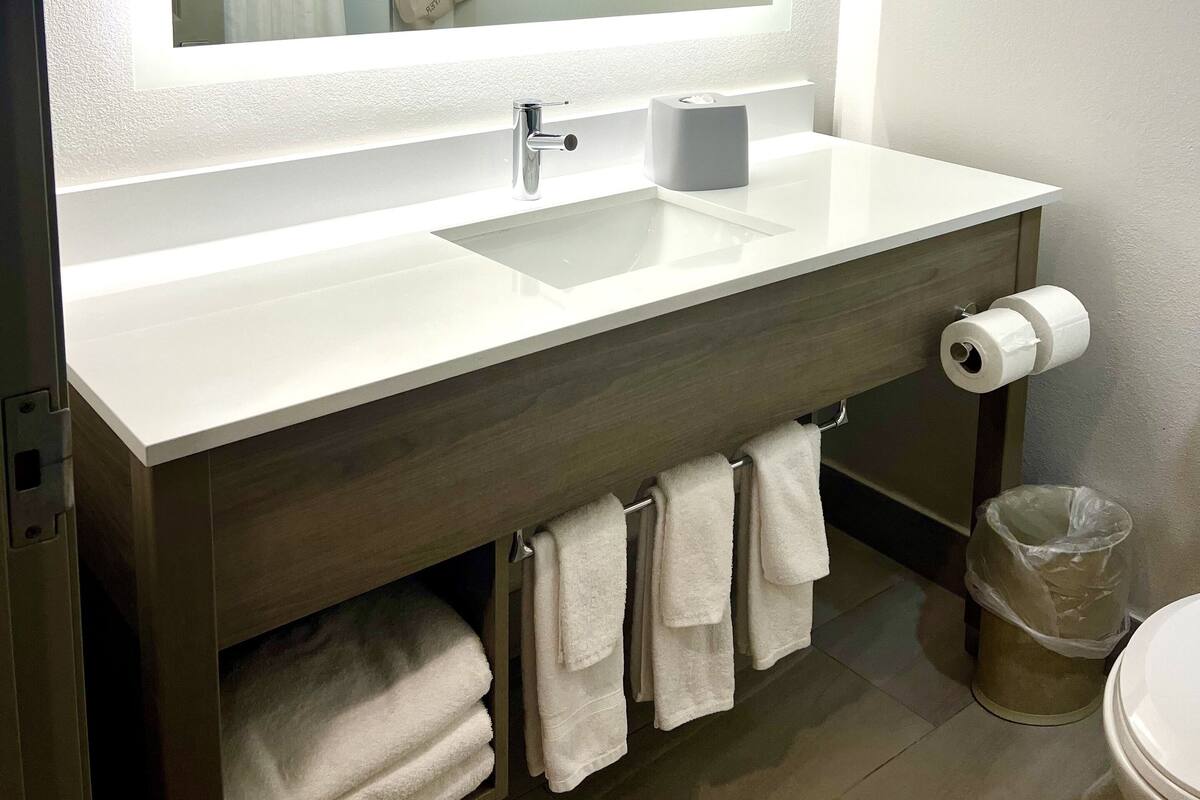 Ein modernes Badezimmer mit einem großen Spiegel, einem weißen Waschbecken, einer Toilette und einem Mülleimer.