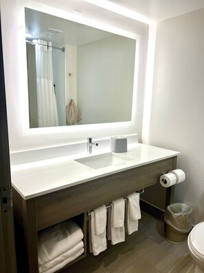 A modern bathroom with a large mirror, a white sink, a toilet, and a trash can.