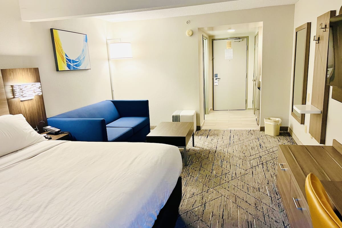Ein Hotelzimmer mit einem Bett, einem blauen Sofa, einem Schreibtisch und einem Stuhl. Ein Flur führt zu einem weiteren Zimmer.