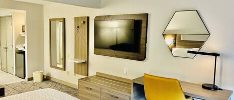 A hotel room with a bed, a desk with a chair, a television, a mirror, and a small cabinet.