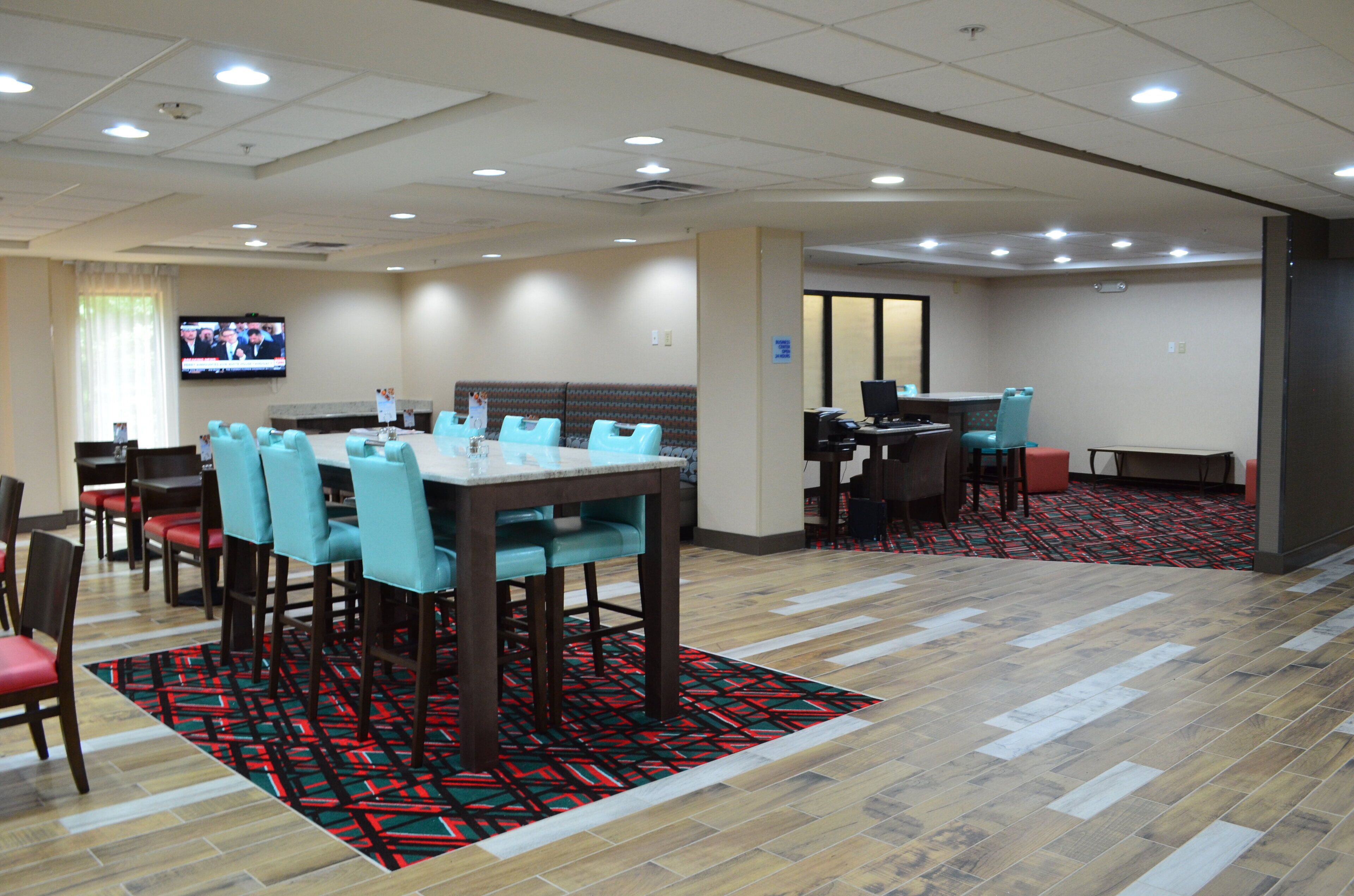 A hotel dining area with a large table and chairs, a television screen, and a patterned carpet.