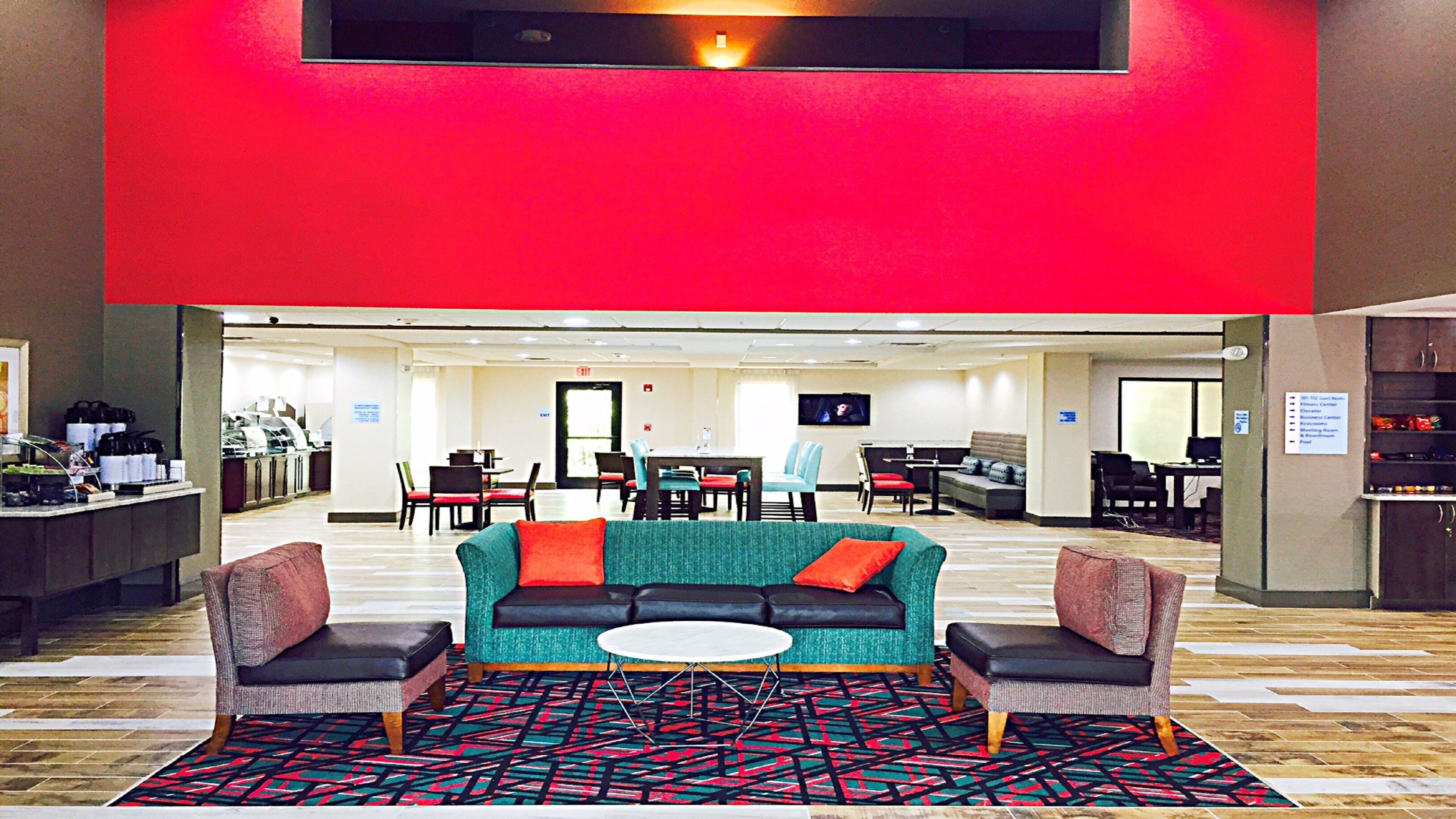 A hotel lobby with a red accent wall, a seating area with a sofa and chairs, a coffee station, and a television screen.