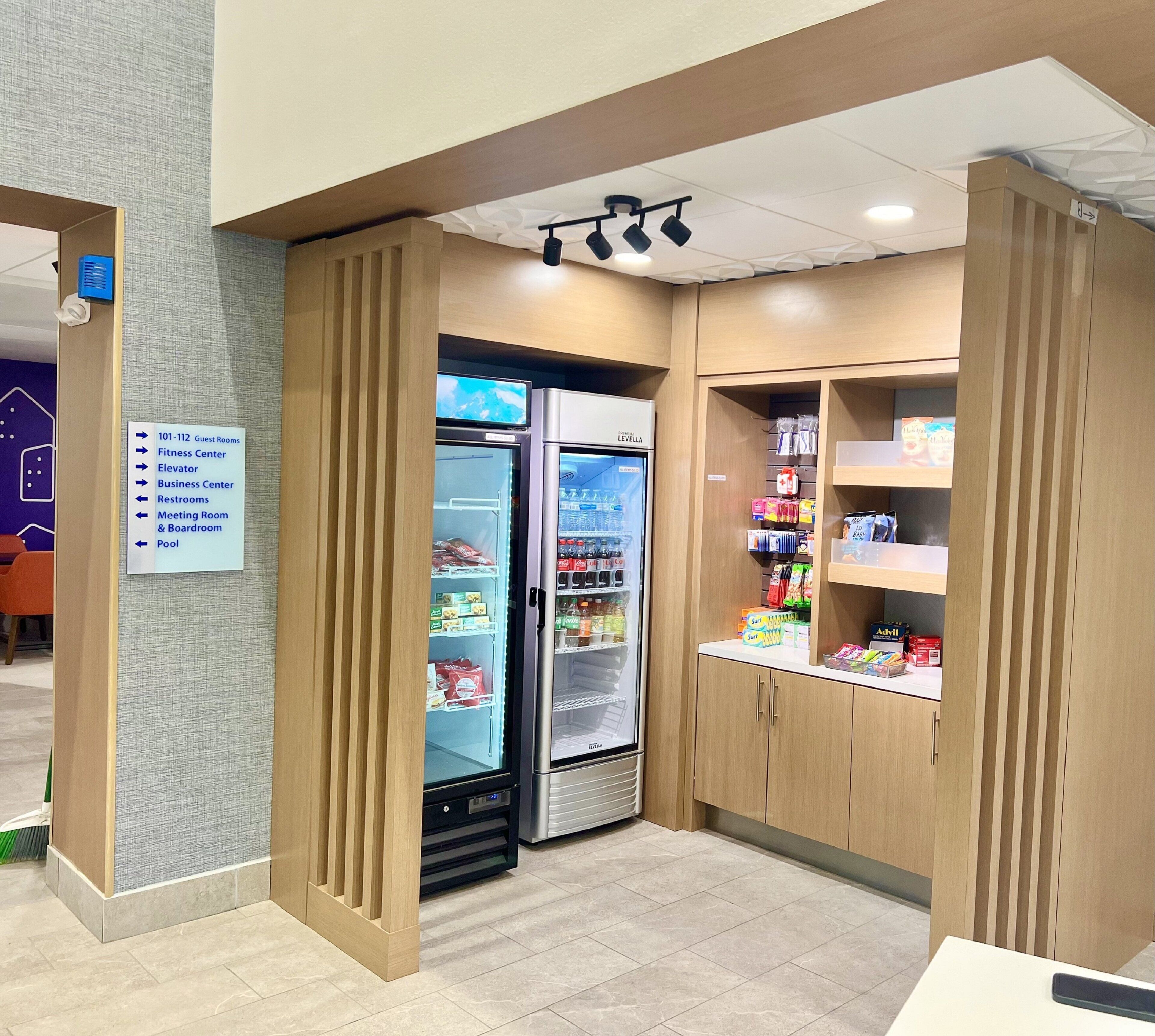 Een hotel lobby met een bord waarop gastenkamers, fitnessruimte, lift, business center, toiletten, vergaderruimte, directeurskamer en zwembad staan aangegeven. Er staan twee koelkasten vol met drankjes en snacks.