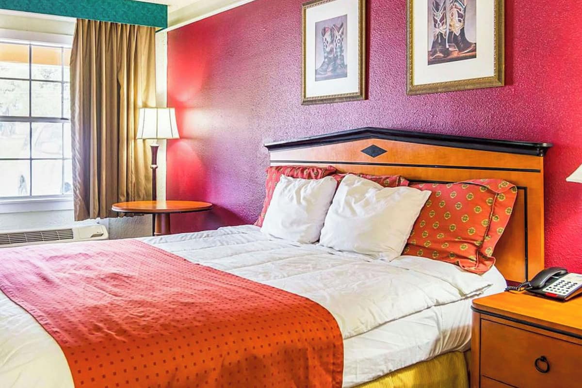 A hotel room with a bed, a bedside table with a phone, a lamp, and a window with curtains.