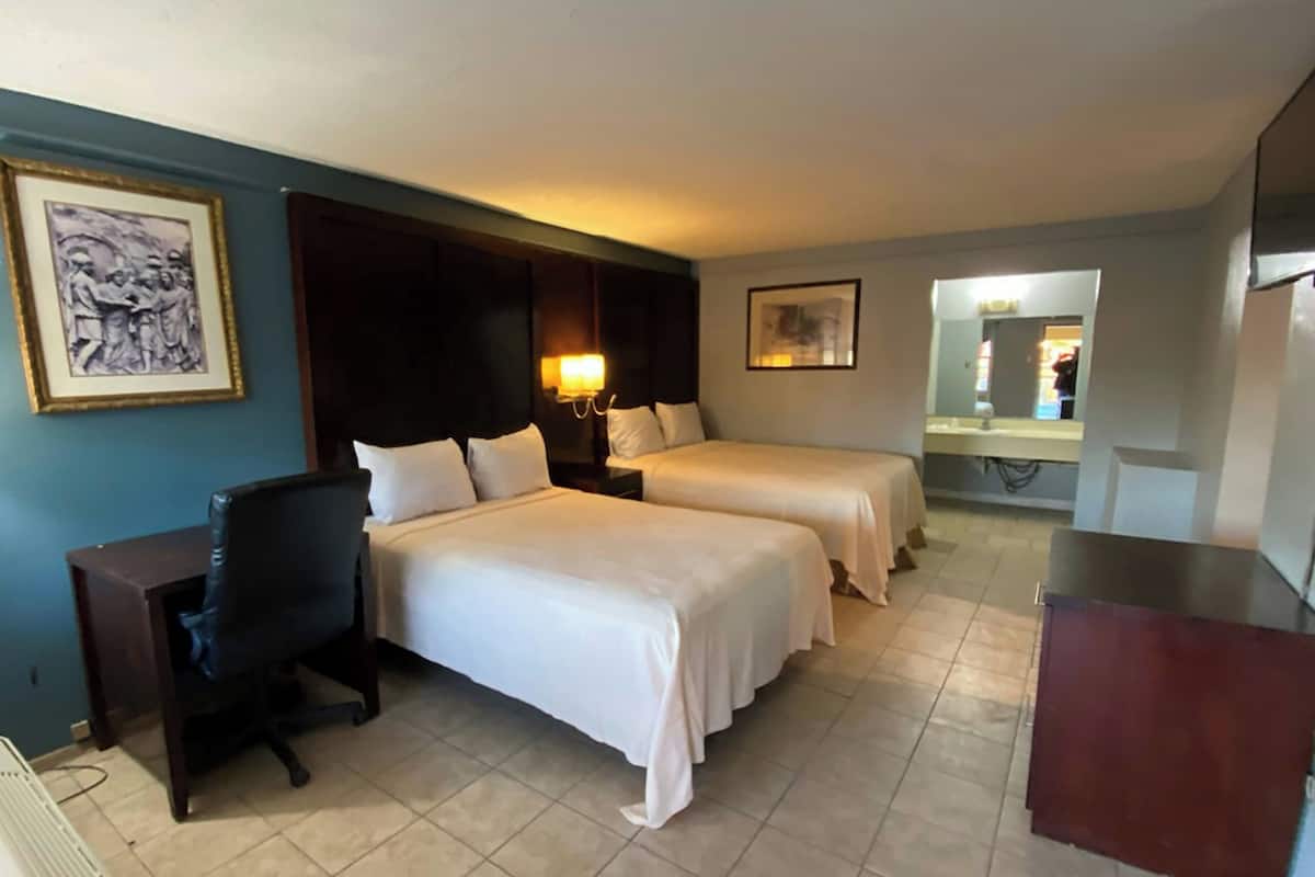 A hotel room with two beds, a desk, a chair, a desk lamp, a framed picture, a mirror, and a TV.