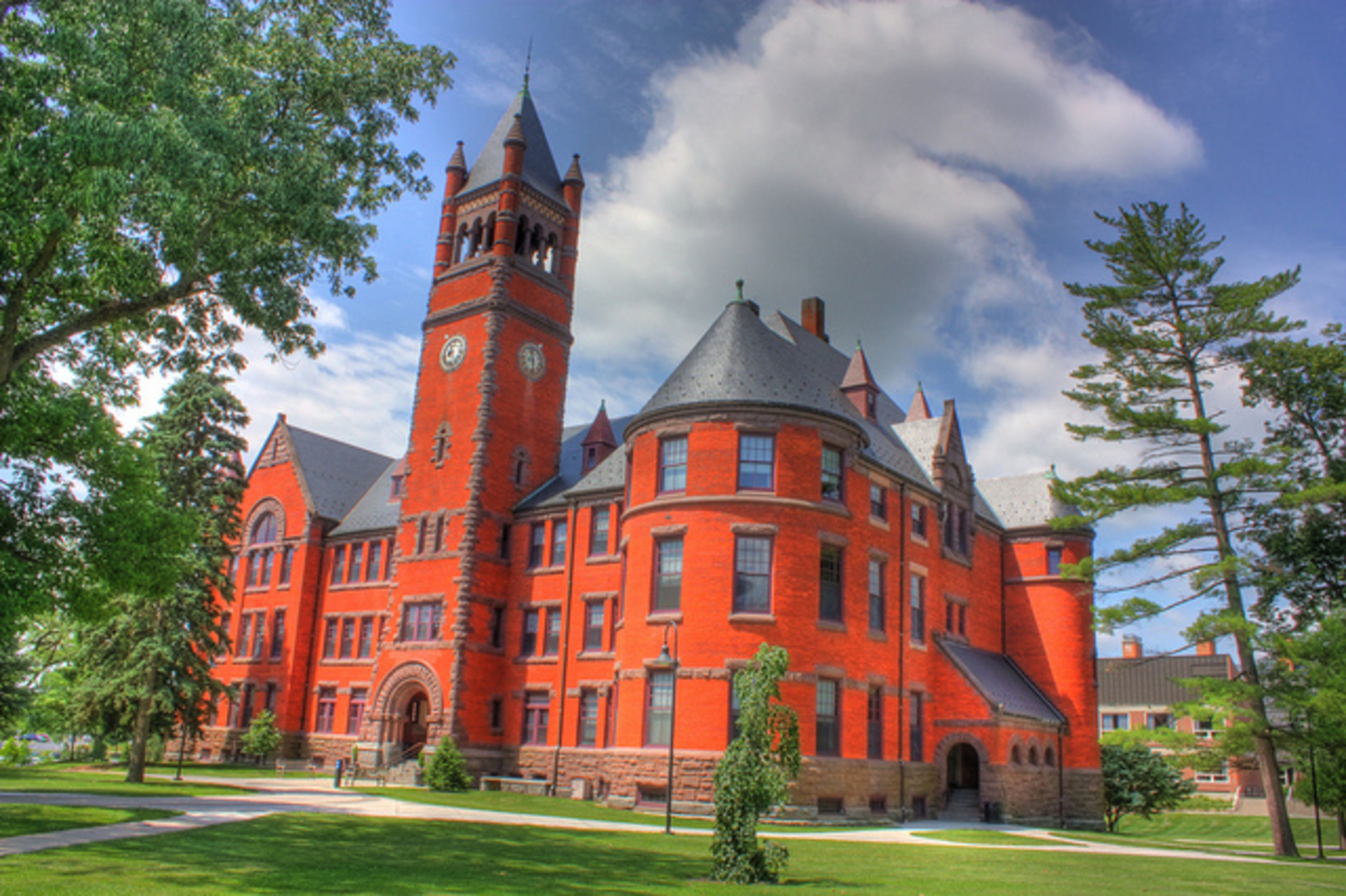 A large, historic red brick building with a prominent clock tower, surrounded by well-maintained lawns and trees.