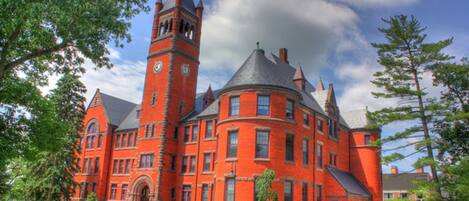 A large, historic red brick building with a prominent clock tower, surrounded by well-maintained lawns and trees.