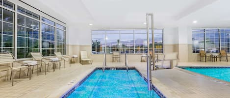 An indoor swimming pool with clear blue water, surrounded by lounge chairs and tables, large windows offering views of the outside.
