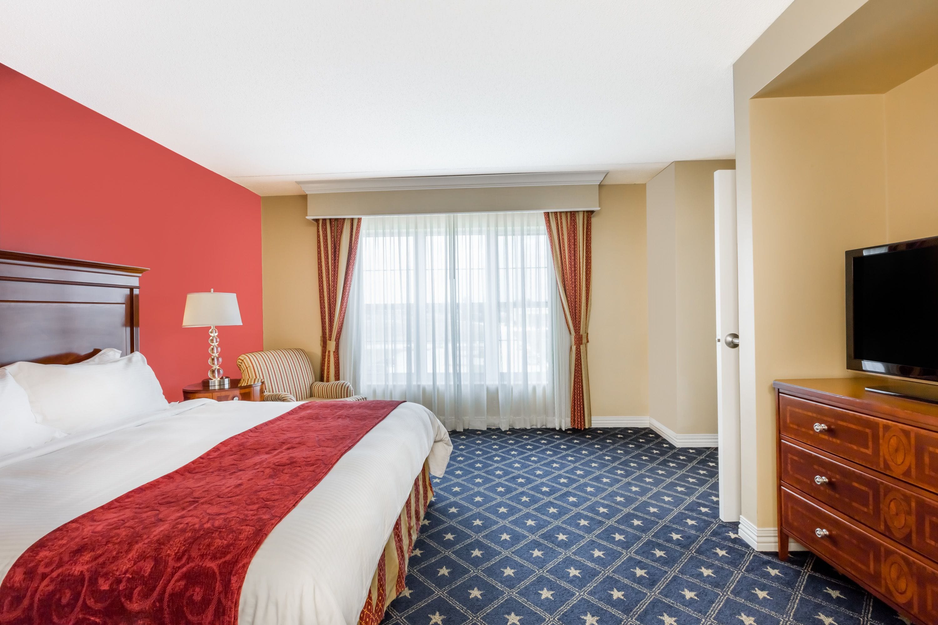 Habitación de hotel con una cama grande, televisión, cómoda y ventana con cortinas.