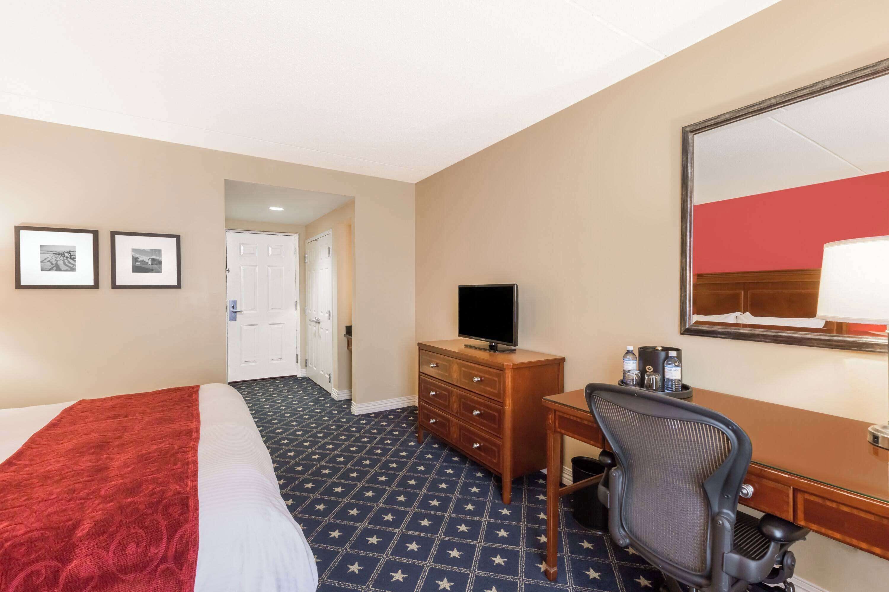 Una habitación de hotel con una cama, un escritorio con una silla, un televisor, un aparador y un espejo.
