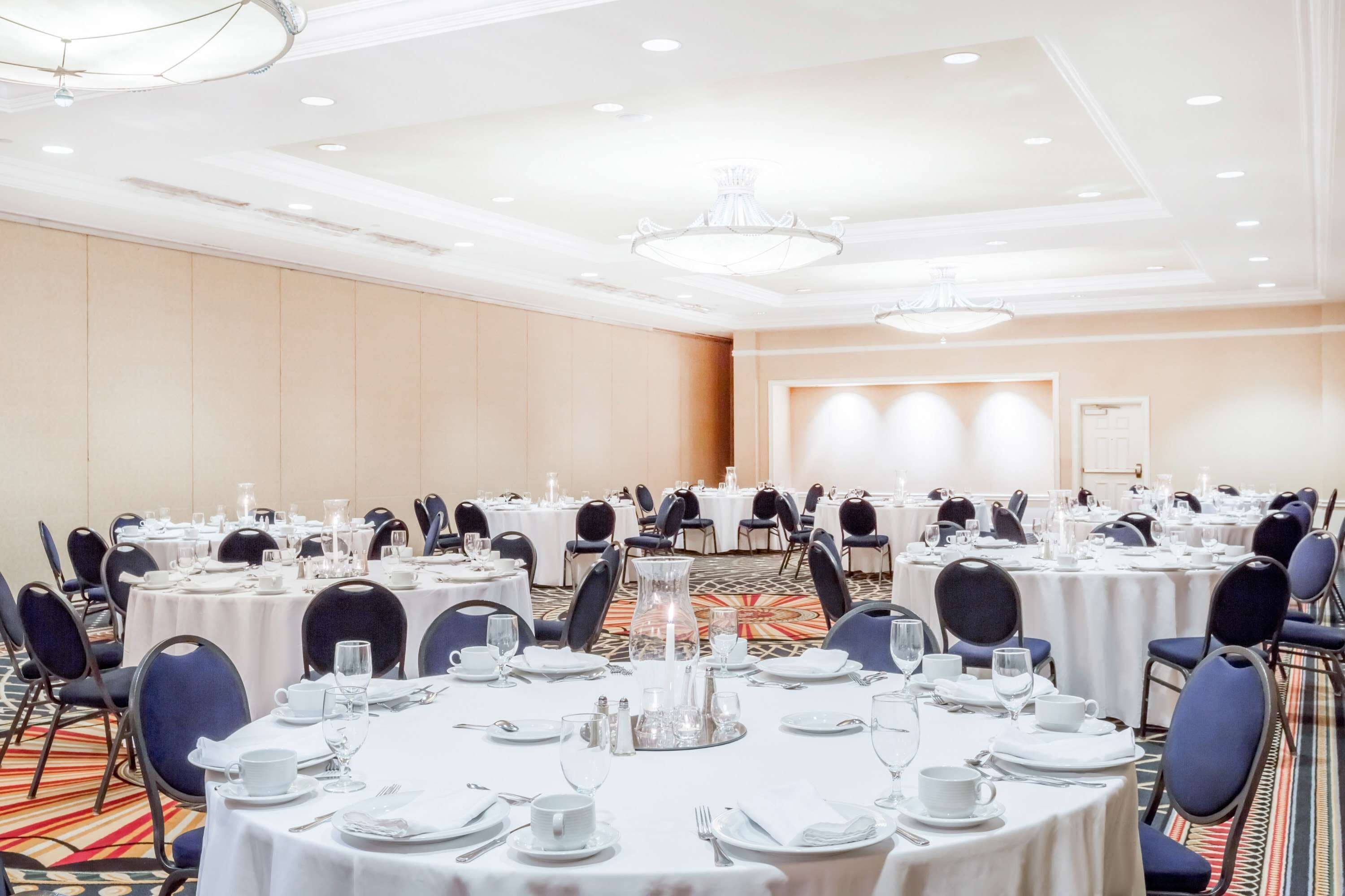 A banquet hall with round tables set for dining, featuring white tablecloths, blue chairs, and a chandelier.