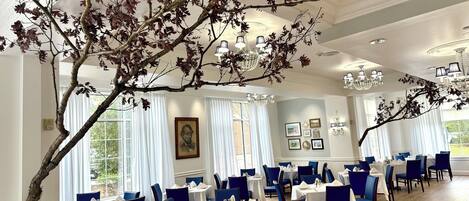 A dining area with multiple tables and chairs, large windows with white curtains, and a chandelier.