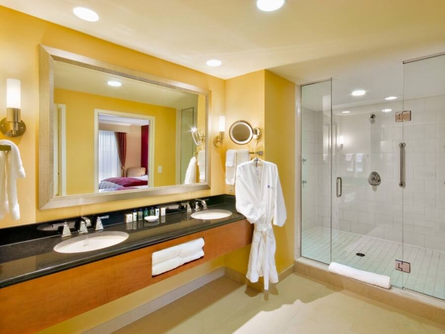 Une salle de bain d’hôtel avec un grand miroir, deux lavabos, une douche en verre et un porte-serviettes.