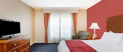 Et hotelværelse med en seng, et fjernsyn på en trækommode, en stol, en lampe og et vindue med gardiner.
