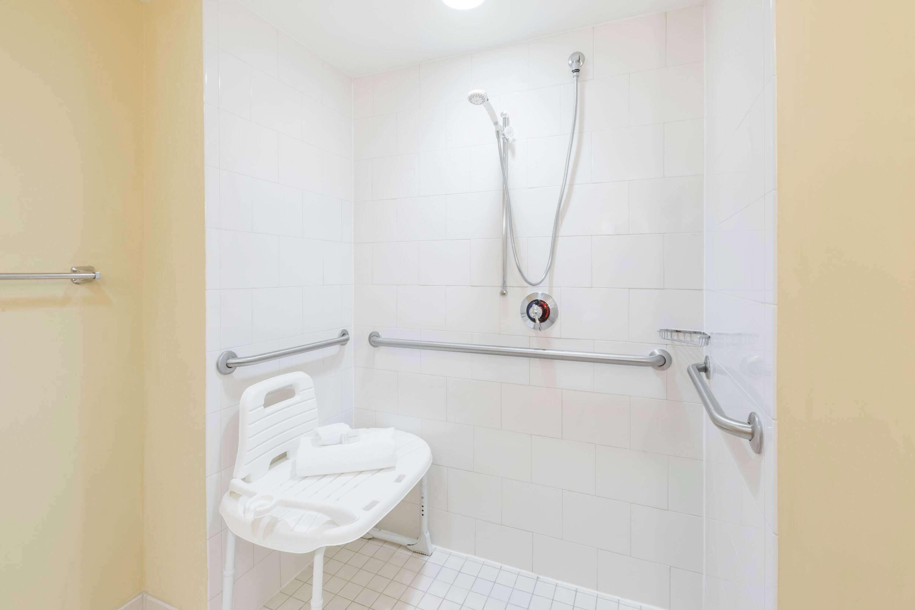 Ein Badezimmer mit weißen Fliesen, einer Dusche inklusive Handbrause und einer weißen Haltestange.