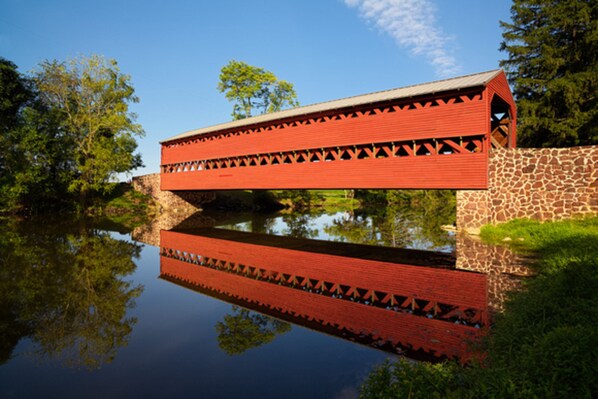 Un pont couvert de couleur rouge, avec un mur de pierre d’un côté et son reflet dans l’eau.
