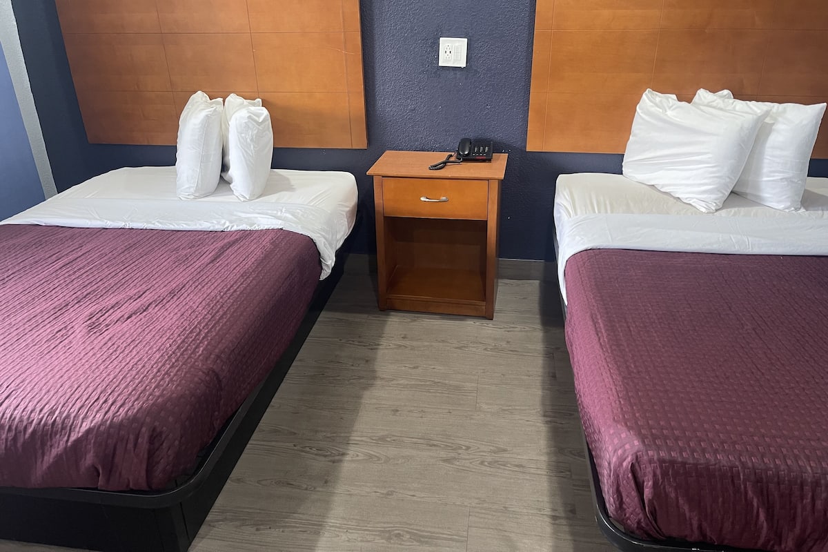 A hotel room with two beds, each with a wooden headboard, a small wooden nightstand with a phone, and a light switch on the wall.