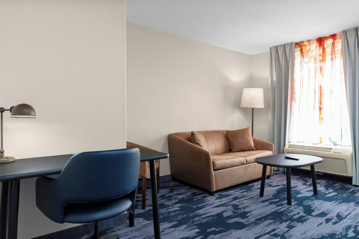 Habitación de hotel con sofá de cuero marrón, un sillón azul, una mesita redonda, un escritorio con lámpara y una ventana con cortinas.