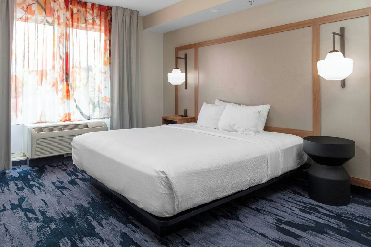 Habitación de hotel con una cama grande, una ventana con cortinas, una mesita de noche y una lámpara fijada a la pared.