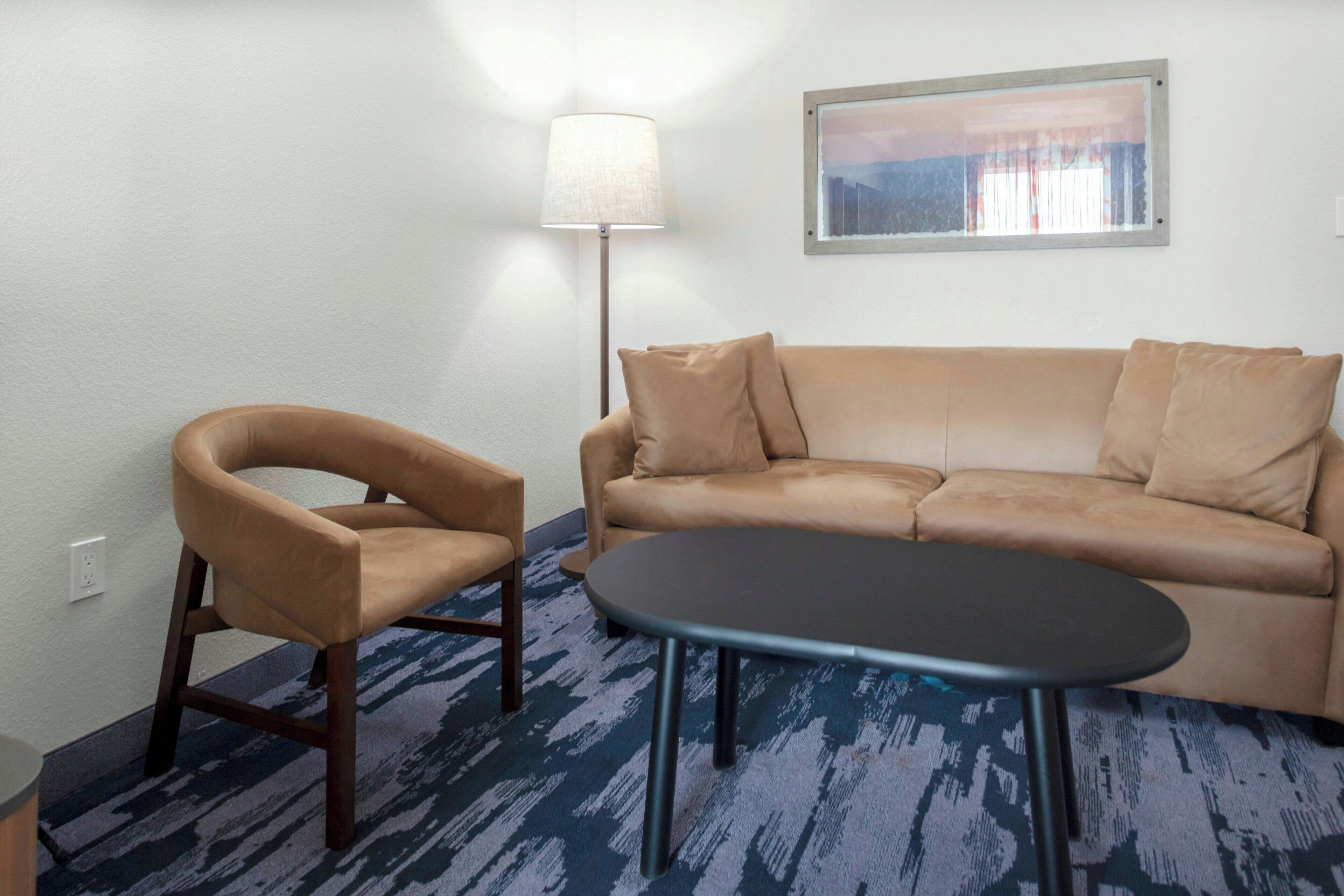 A living room with a beige sofa, a single armchair, a round coffee table, a floor lamp, and a framed picture on the wall.