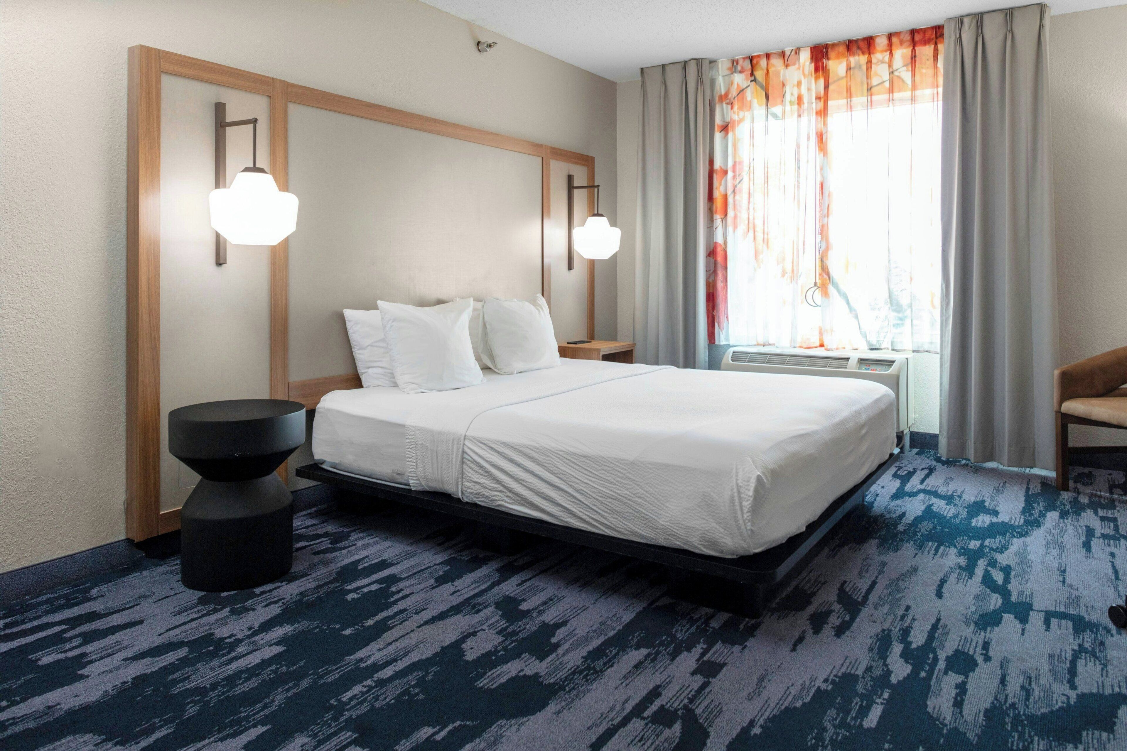 Ein Hotelzimmer mit einem großen Bett, zwei Nachttischen, einem runden schwarzen Beistelltisch, einem Fenster mit Vorhängen und einem Sessel.