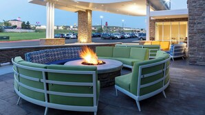 A patio area with a fire pit, green cushioned seating, and a view of a parking lot and buildings in the background.