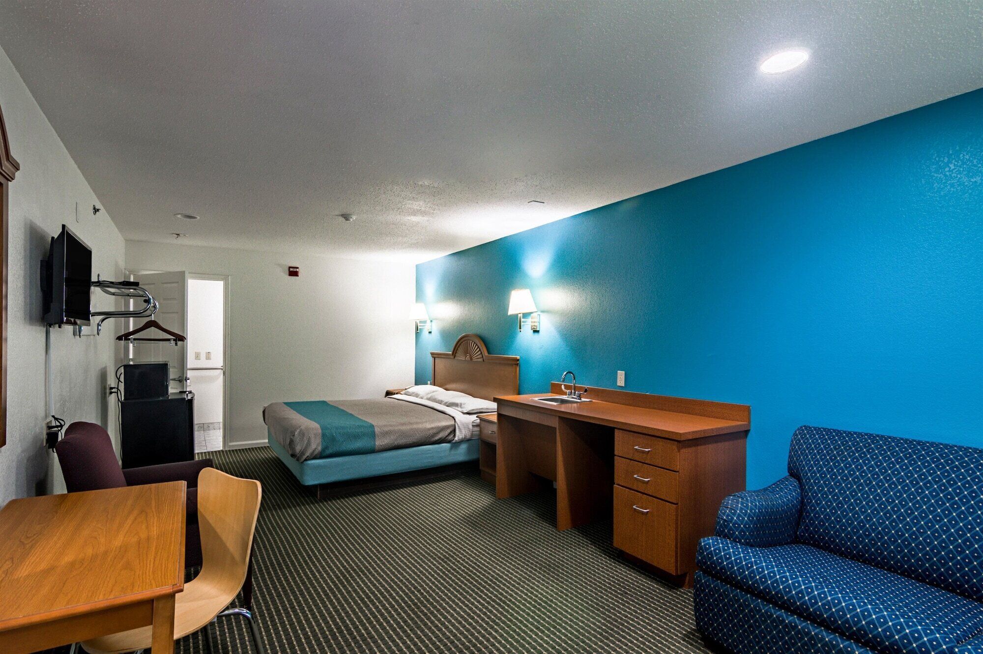 A hotel room with a bed, a desk with a chair, a sofa, a TV mounted on the wall, and a bathroom with a mirror and sink.