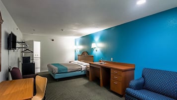 A hotel room with a bed, a desk with a chair, a sofa, a TV mounted on the wall, and a bathroom with a mirror and sink.