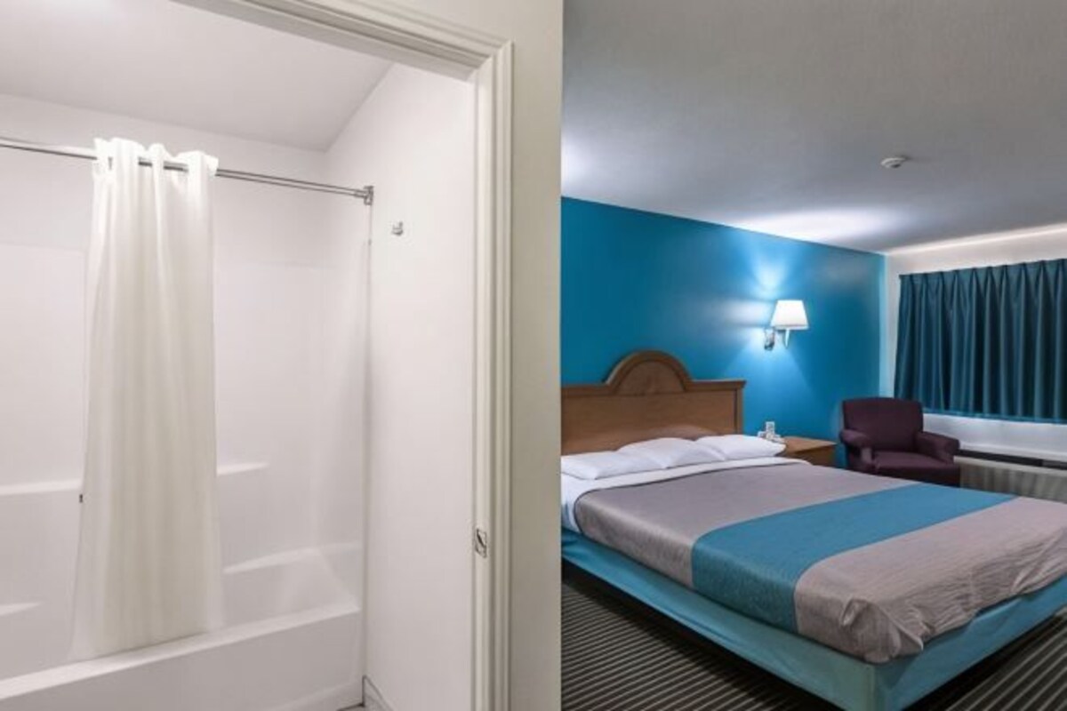 A hotel room with a shower, a bed with a headboard, a nightstand, a chair, and a window with curtains.