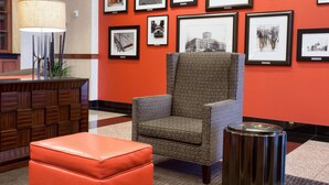 A lobby with a patterned armchair, a red ottoman, a lamp, and a wall adorned with framed photographs.