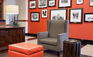 Eine Lobby mit Sesseln mit Muster, einem roten Hocker, einer Lampe und gerahmten Fotos an der Wand.