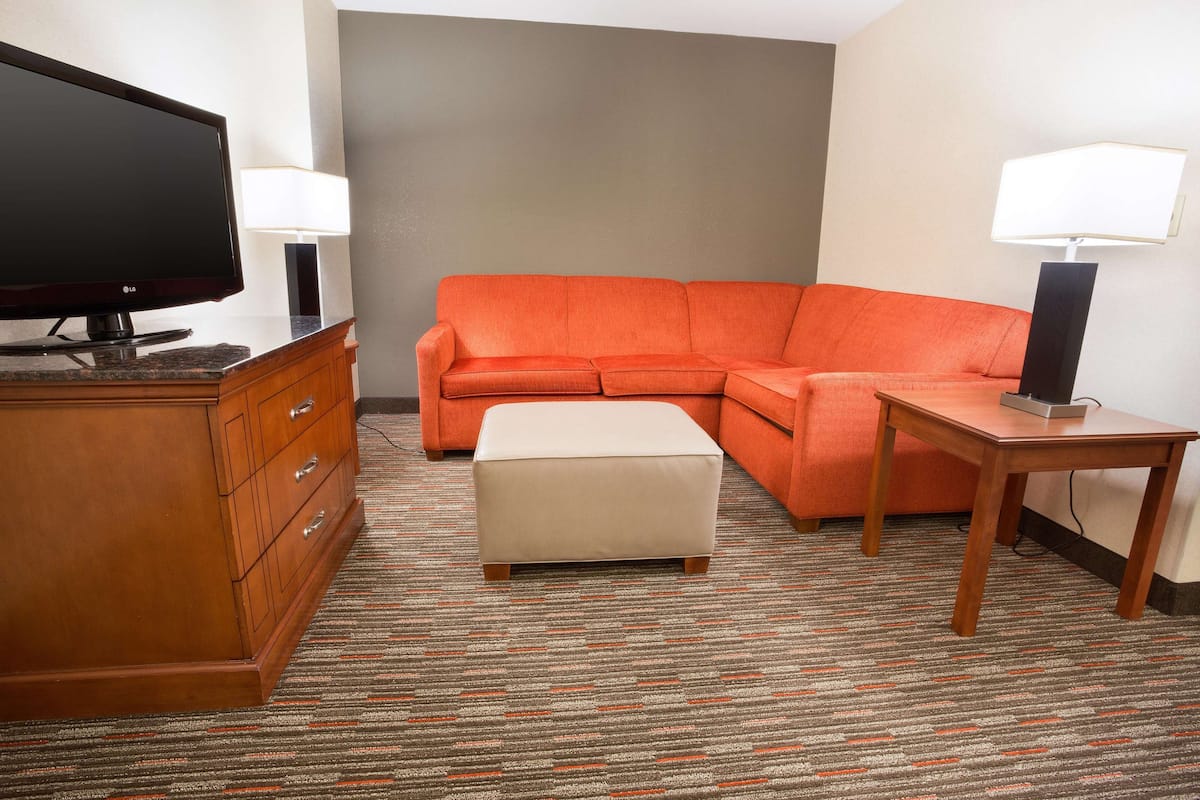 Ein Hotelzimmer mit einem Holzkoffer, einem Fernseher, einem orangefarbenen Sofa, einem passenden Hocker und einem kleinen Holztisch mit einer Lampe.