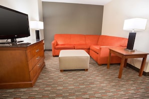 Ein Hotelzimmer mit einem Holzkoffer, einem Fernseher, einem orangefarbenen Sofa, einem passenden Hocker und einem kleinen Holztisch mit einer Lampe.