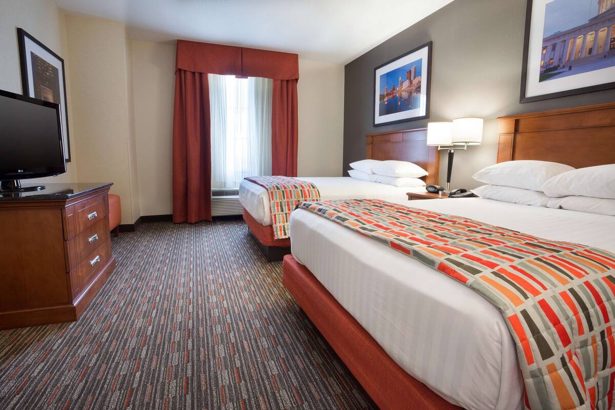 Ein Hotelzimmer mit zwei Betten, einem Fernseher, einem Schreibtisch und einem Fenster mit Vorhängen.