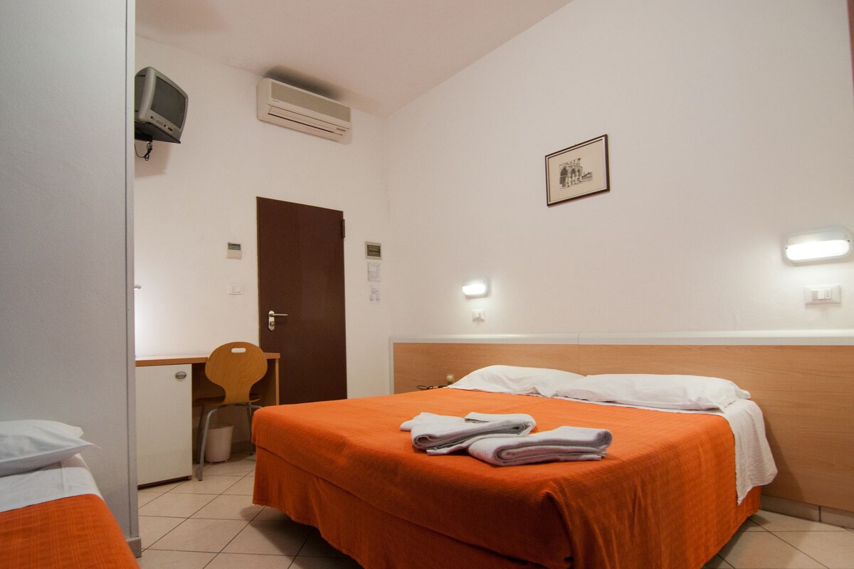 A hotel room with a bed, a desk, and a chair. There is a door, an air conditioner, and a framed picture on the wall.